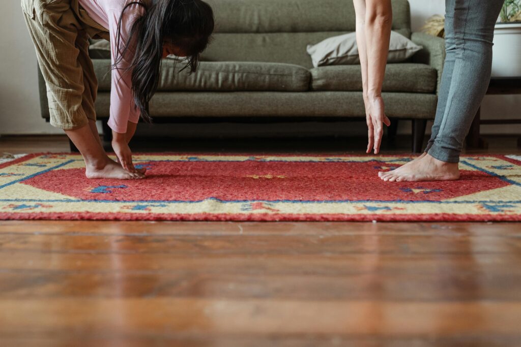 Side view of young ethnic woman stretching body with adorable girl while standing against each other on floor carpet near cozy sofa in modern living room in morning